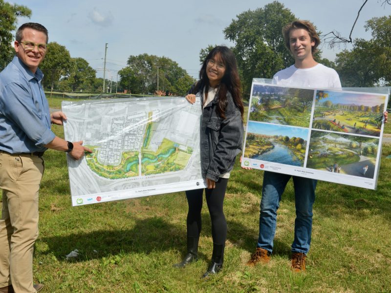 Three people stand in a field holding posterboard that depicts a landscape improvement project.