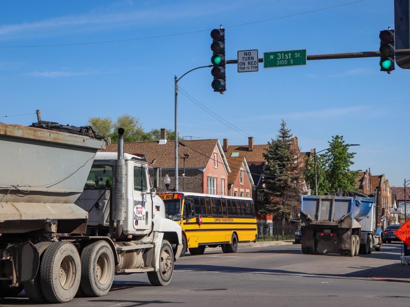 Two empty dump trucks drive northbound on Pulaski while a school bus drives south. A pedestrian can be seen walking south on Pulaski Rd. The 31st St. street sign is visible.