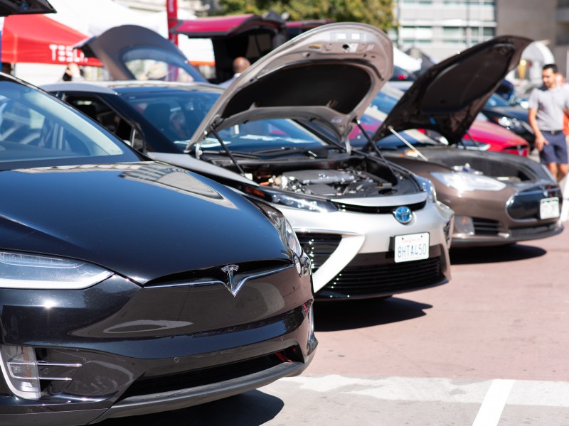 Three electric vehicles are parked in a line, and two have their hoods open.