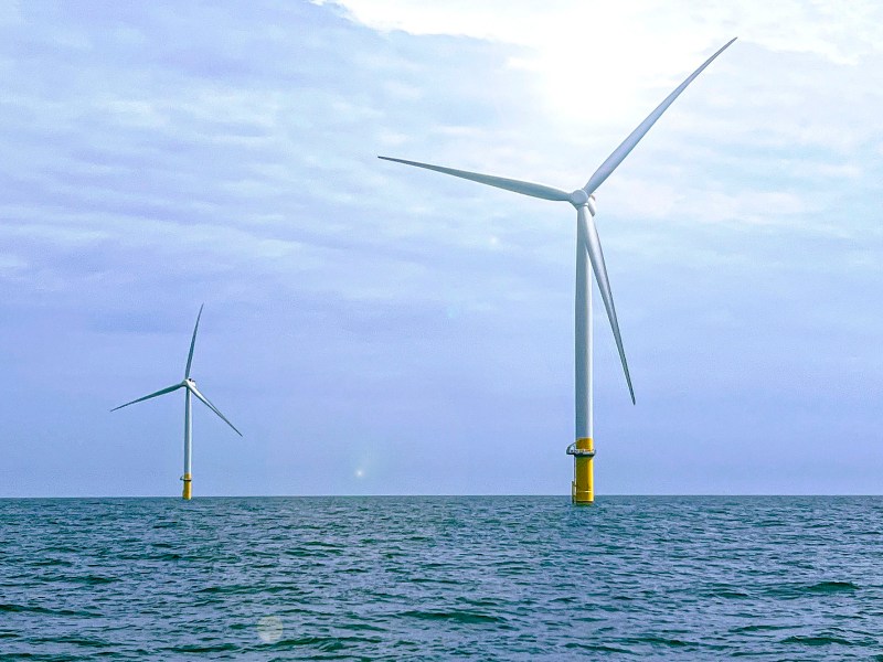 One wind turbine stands in ocean against a cloudy sky in the foreground, and another stands in the back.