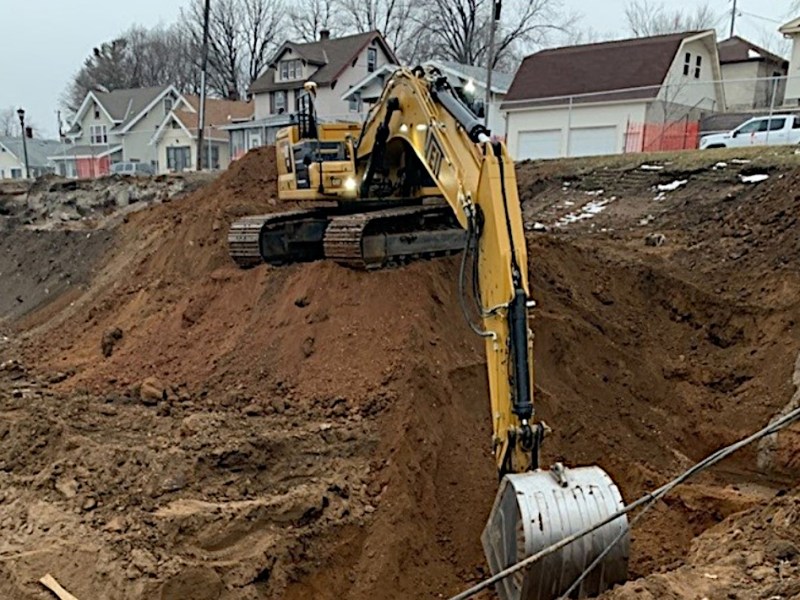 An excavator digs a hole in a schoolyard along a residential street.