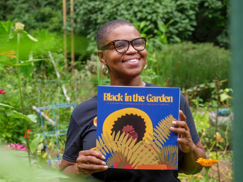 Colah B Tawkin stands in a garden with a copy of a coloring book bearing the name of her podcast, "Black in the Garden"