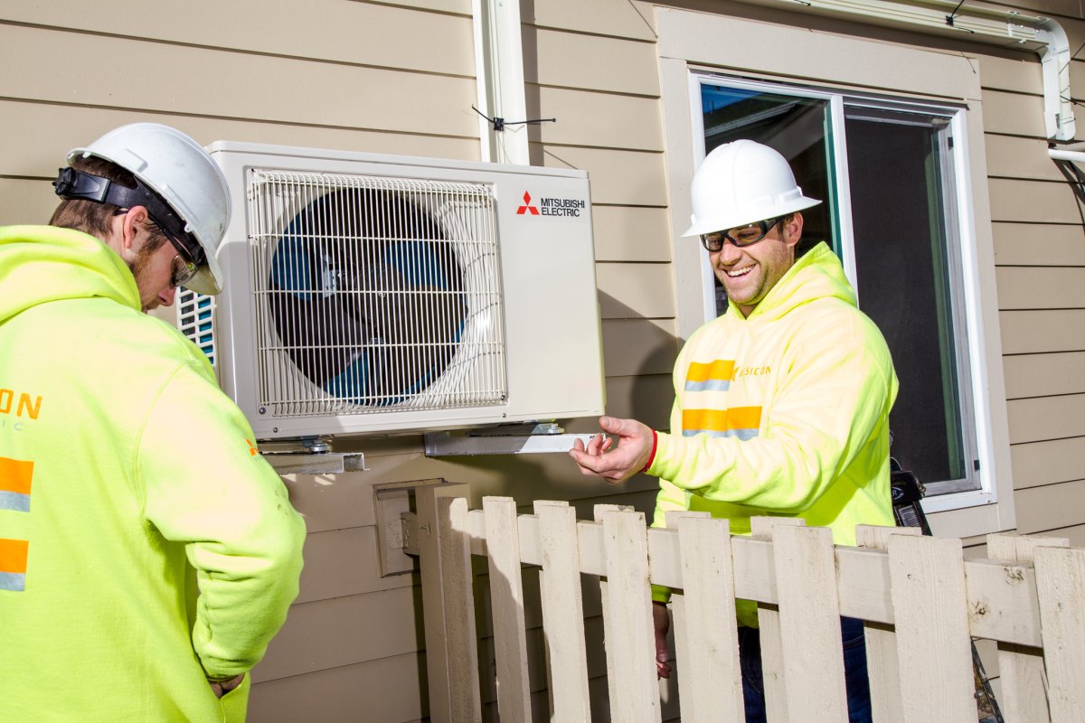 Jovial workers in hard hats installing a heat pump on the side of a house.