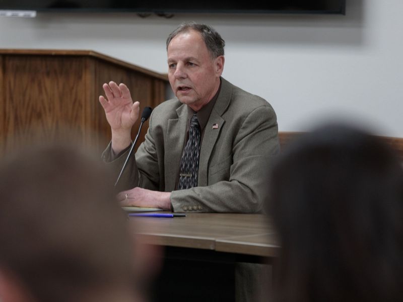 A man in a grey suit sits at a table speaking before a crowd.