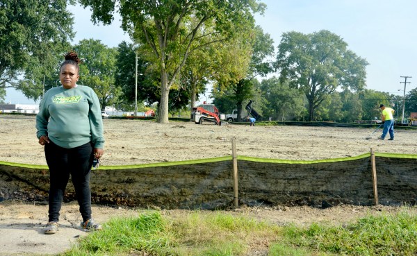 A woman in a green sweatshirt stands in front of an open dirt patch where someone is working. It's surrounded by a low black plastic fence.