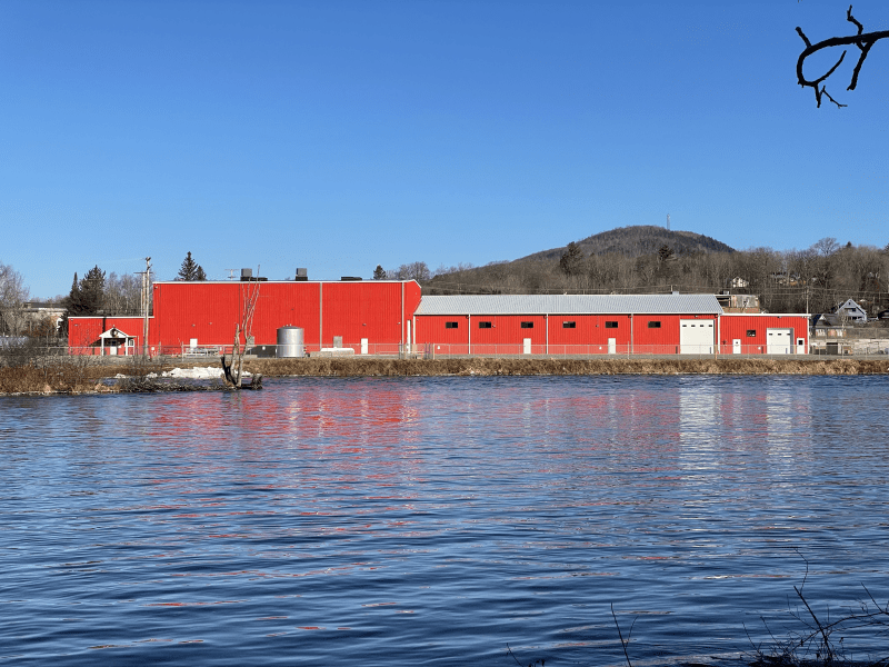 A long red building stands on the shore of a river, with a blue sky above.