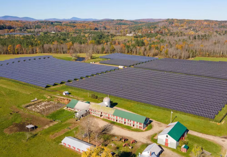 A large solar array on a field next to a dairy farm, with hills in the background.